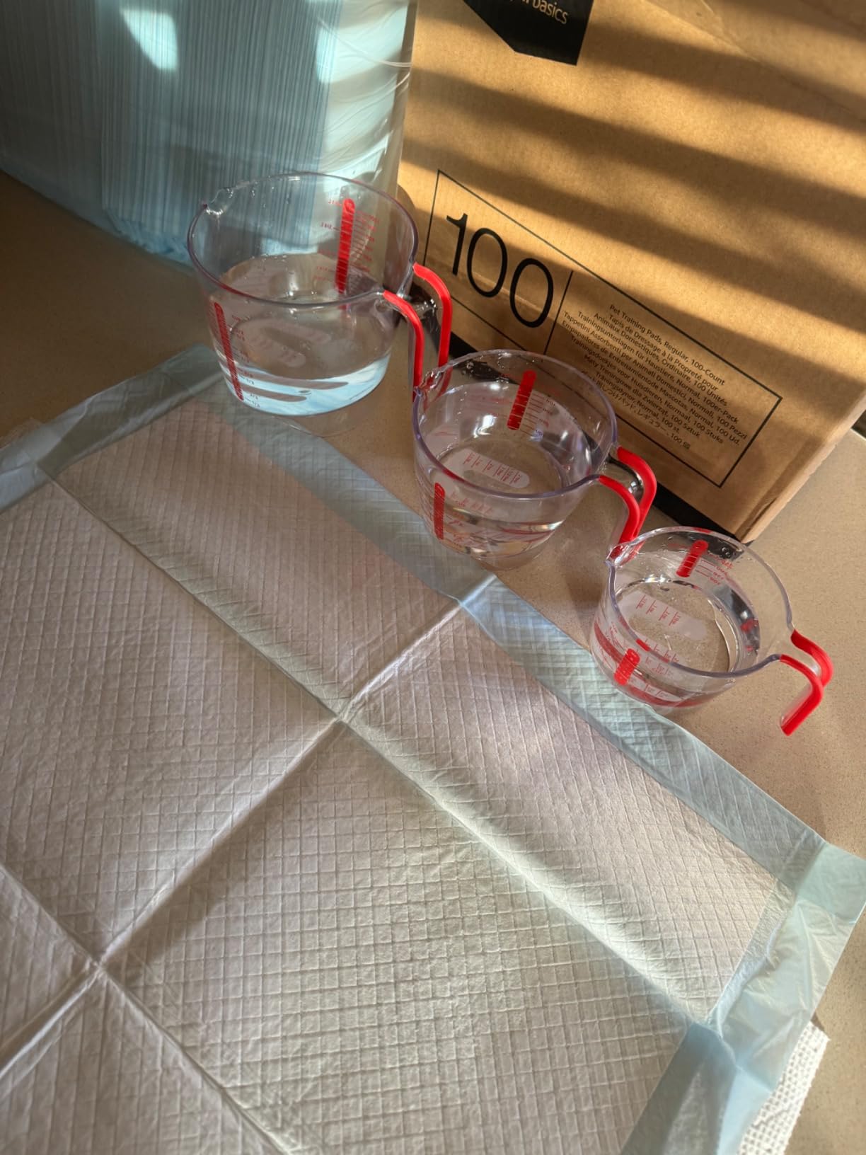 Customer absorbency test with three glass measuring cups of water poured onto one blue backed training pad on a kitchen floor, next to the 100 count box it came in
