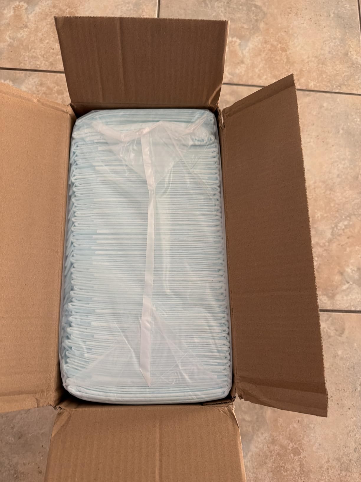 Opened shipping box on a tiled floor with a shrink-wrapped stack of pale blue disposable underpads filling it
