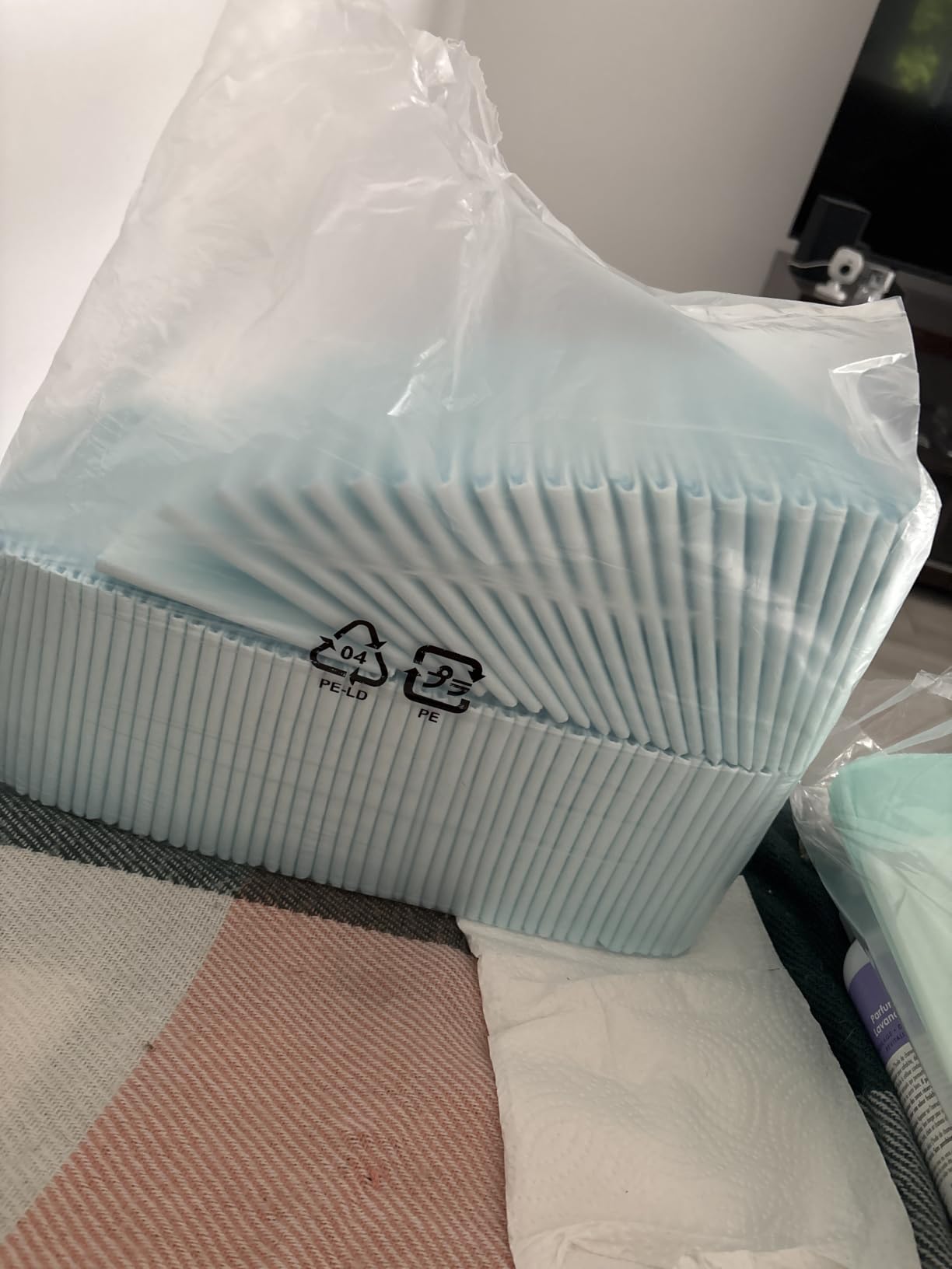 Close view of a plastic-wrapped stack of blue disposable underpads resting on a bed, with the recycling marks printed on the packaging visible