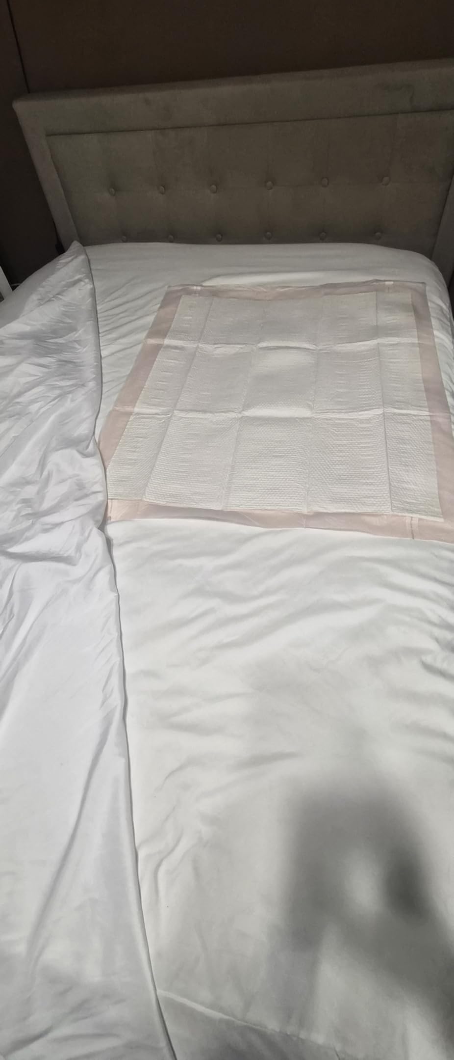 One peach backed underpad laid flat on a white bed below a tufted headboard, with the sheet folded back over it
