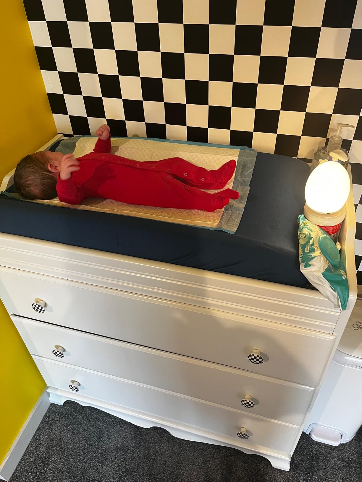 Newborn in a red sleepsuit lying on a blue backed disposable underpad on top of a changing mat, with wipes and a lamp beside it