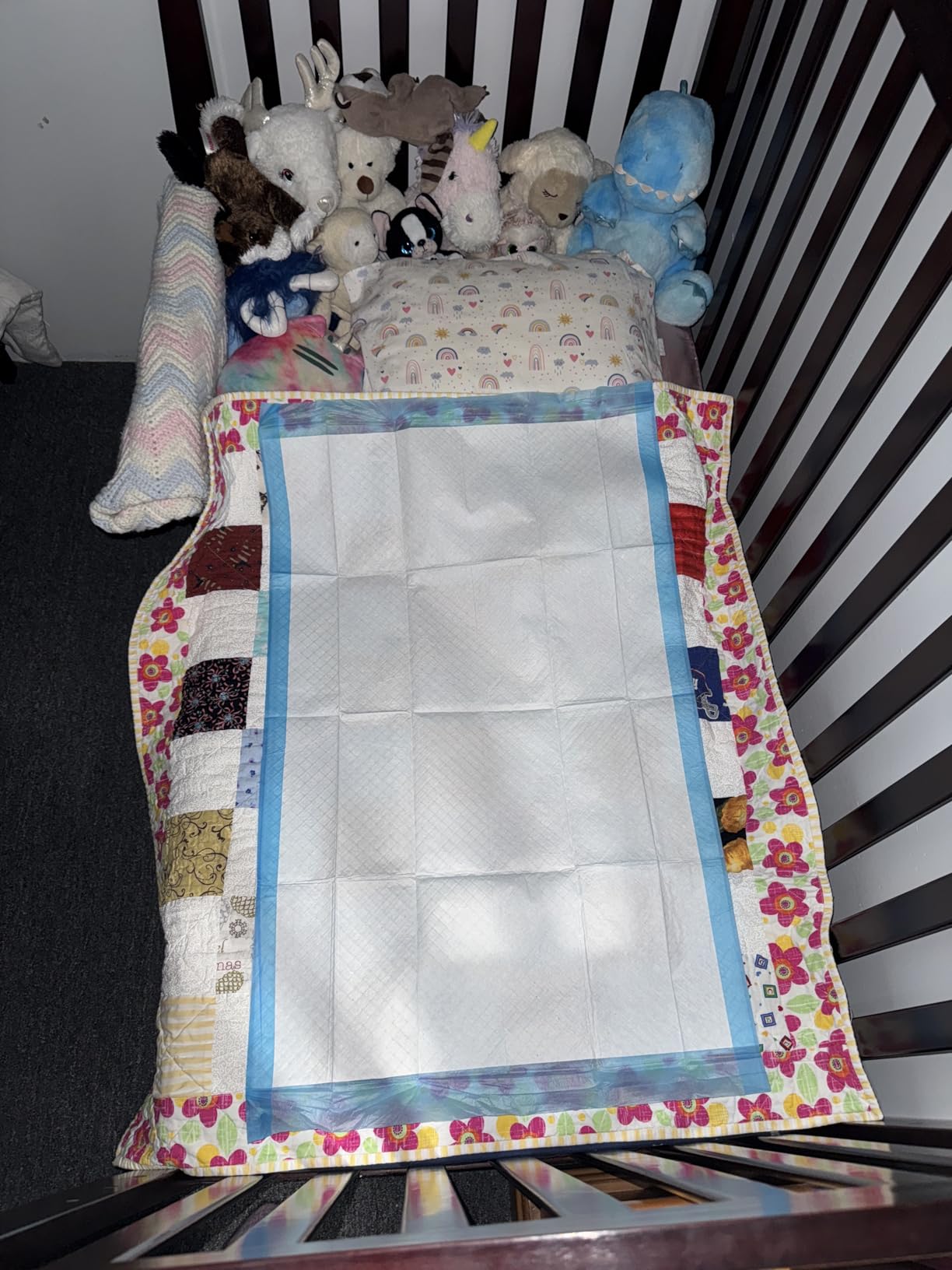Blue backed underpad spread over a patchwork quilt inside a crib, with soft toys stacked at the head end
